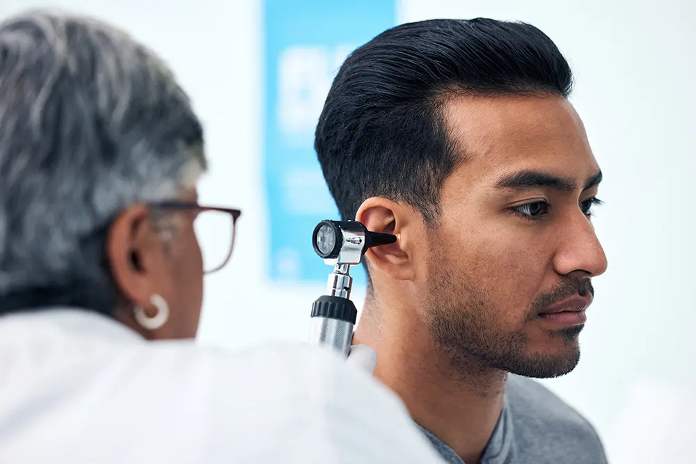 A person is having their ear examined by a healthcare professional using an otoscope. The setting appears to be a medical clinic, and the healthcare professional is wearing glasses and a white coat.