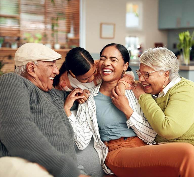 Three adults and one child sit close together on a couch, laughing and hugging. The scene is warm and joyful, in a cozy, sunlit living room.