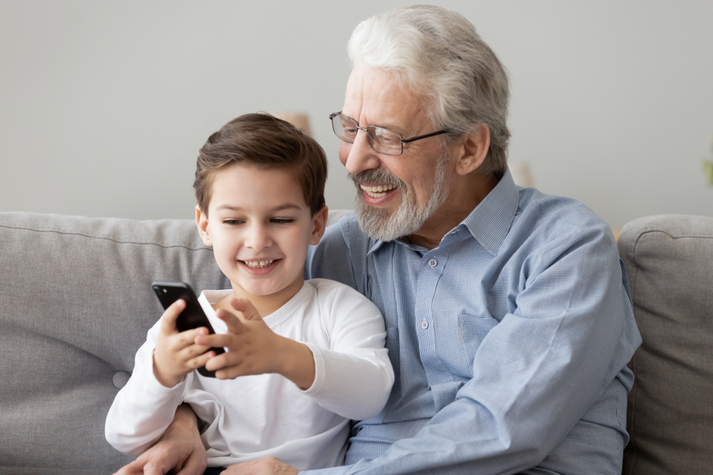 Smiling elderly man with gray hair and beard sitting on a couch with a young boy. The boy is holding a smartphone and showing it to the man, and they both look happy and engaged.