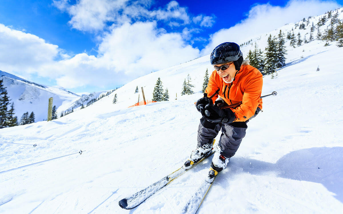 A person wearing an orange jacket, black helmet, and goggles skis downhill on a snowy mountain, smiling with trees and a bright blue sky in the background.