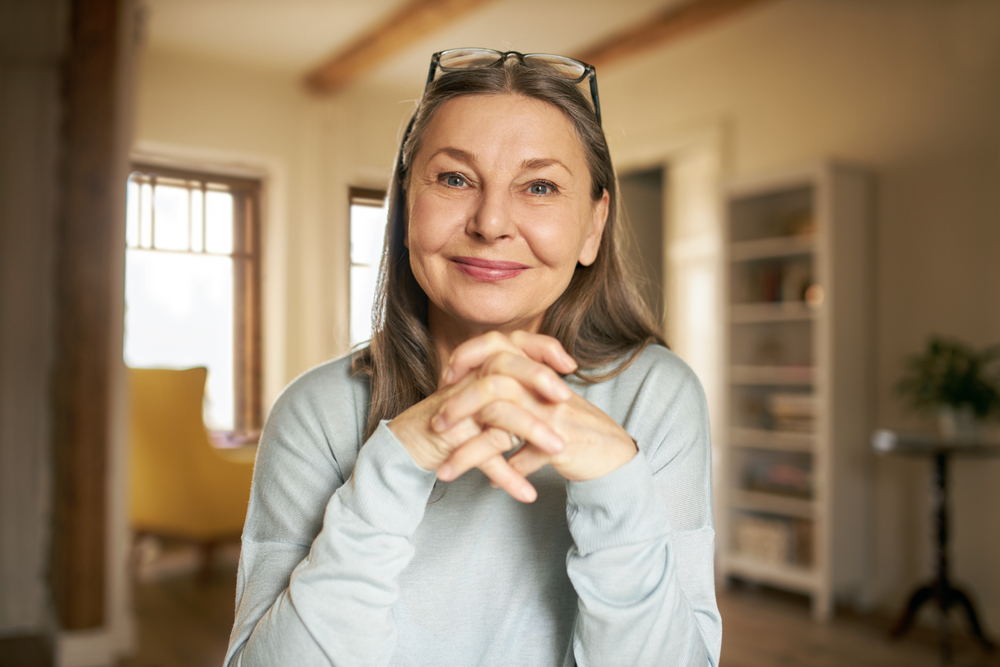 A smiling older woman with long gray hair, wearing glasses on her head and a light blue sweater, sits indoors with her hands clasped, in a warmly lit room with a bookcase and yellow chair in the background.