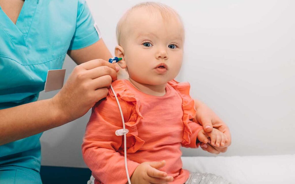 A healthcare professional in scrubs tests a baby’s hearing by placing a small device in the baby’s ear. The baby, wearing a pink shirt with ruffles, looks ahead while sitting on an exam table.