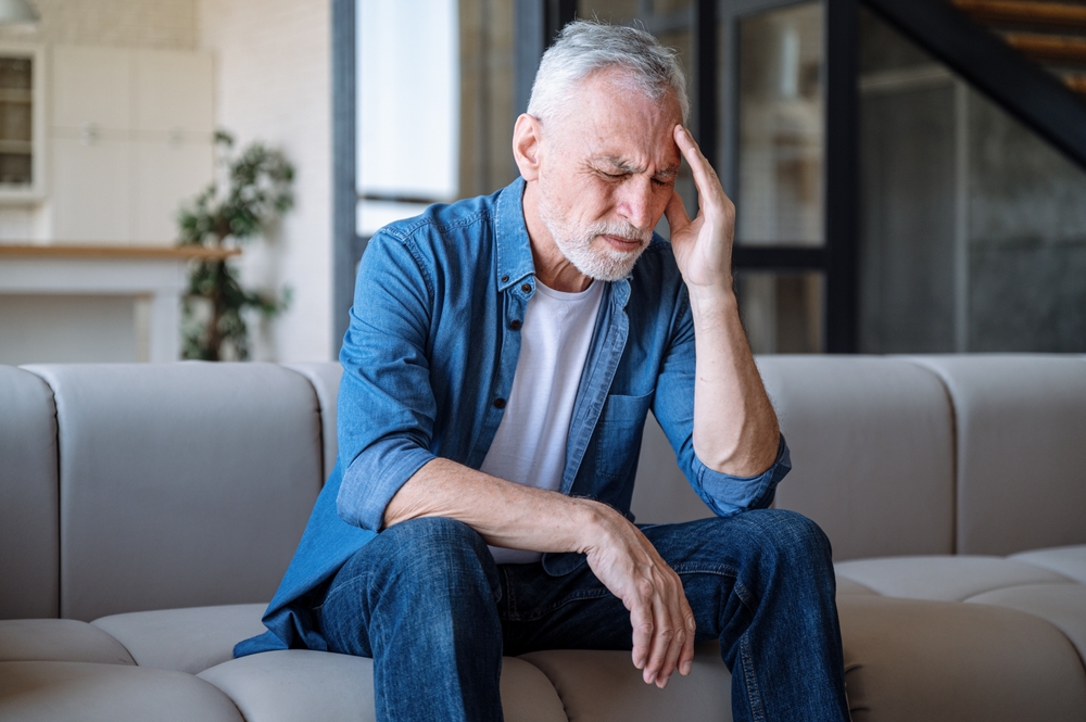 An older man with gray hair sits on a couch, touching his forehead with a pained expression, suggesting he has a headache or is feeling unwell. He wears a denim shirt and jeans in a modern living room.
