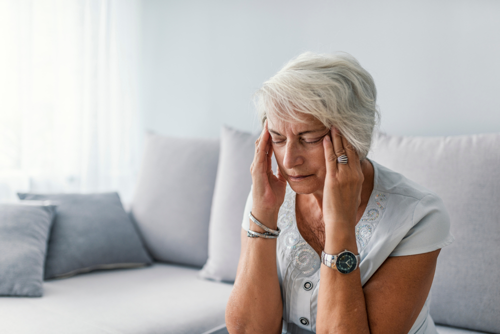 An older woman with short gray hair sits on a couch, eyes closed, touching her temples with both hands, appearing to be in pain or stressed.