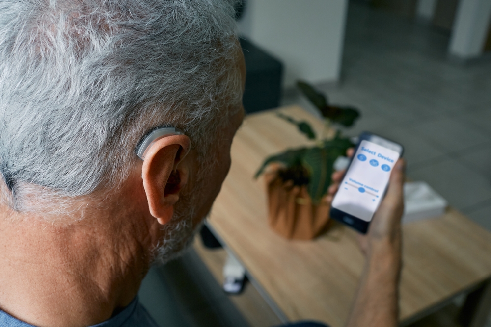 A man with gray hair wears a hearing aid and holds a smartphone, adjusting settings for his device via an app. A table with a potted plant is visible in the background.
