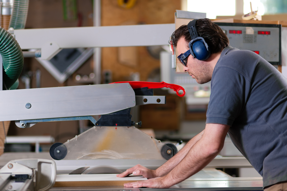 A man wearing safety glasses and earmuffs operates a table saw in a workshop, guiding a piece of wood through the blade with both hands.