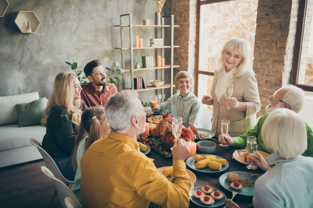 A smiling elderly woman gives a toast at a dining table surrounded by family members of various ages, sharing a festive meal with turkey, corn, and desserts in a bright, cozy room.