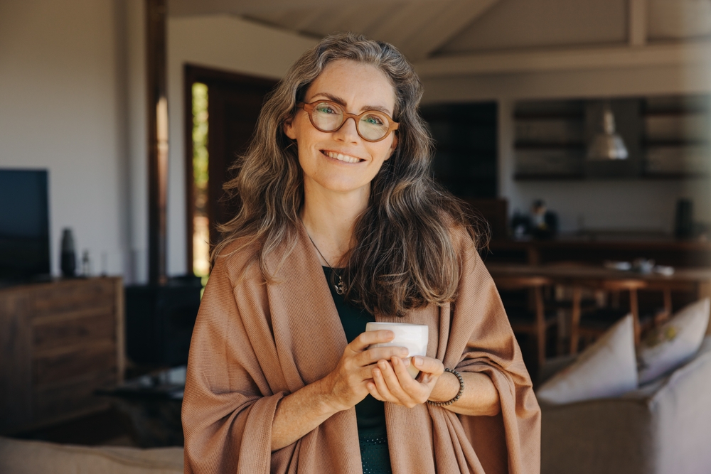 A smiling woman with long gray hair and glasses stands indoors, wearing a brown shawl and holding a white mug. The background shows a cozy, modern living and dining area with wooden furniture.