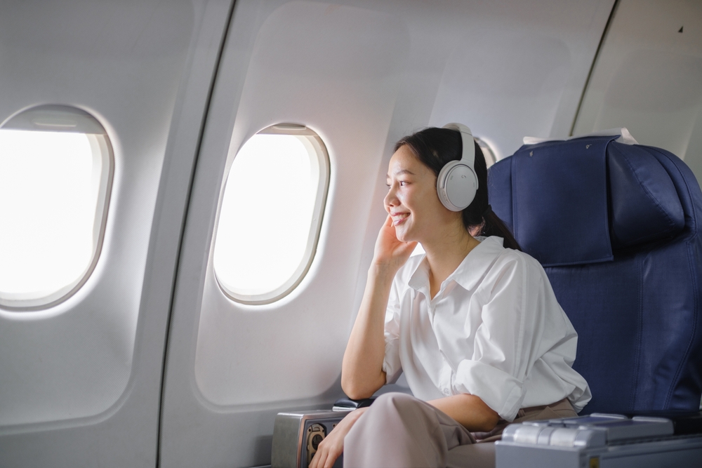 A woman wearing headphones sits in an airplane seat by the window, smiling and looking outside during a flight. She appears relaxed and content, dressed in a white shirt and beige pants.