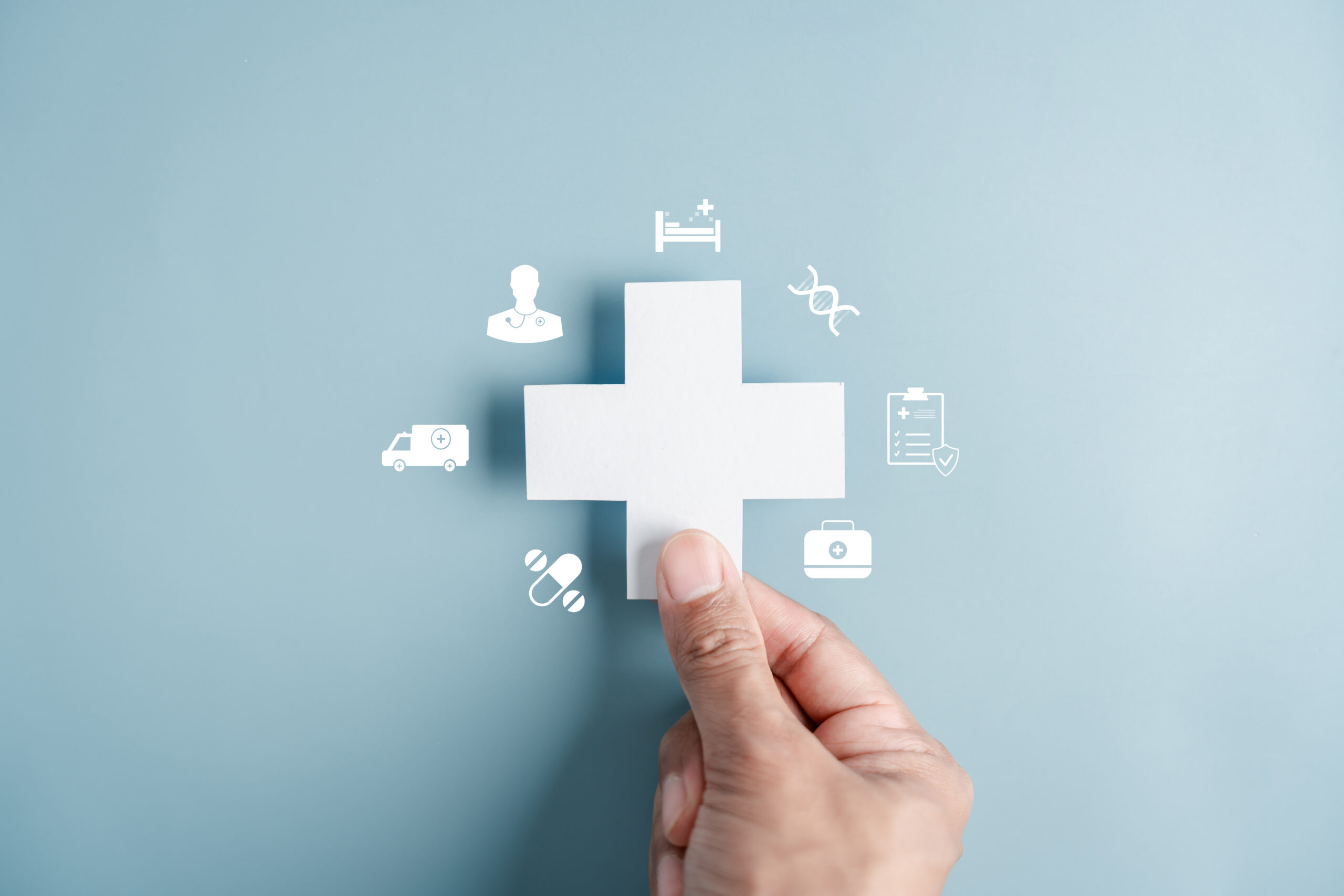 A hand holds a white medical cross on a blue background, surrounded by healthcare icons including a hospital bed, ambulance, pills, a doctor, DNA, a clipboard, and a laptop.