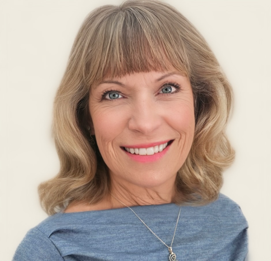 Smiling middle-aged woman with shoulder-length blonde hair and blue eyes, wearing a blue top and a silver necklace, posed against a plain light background.